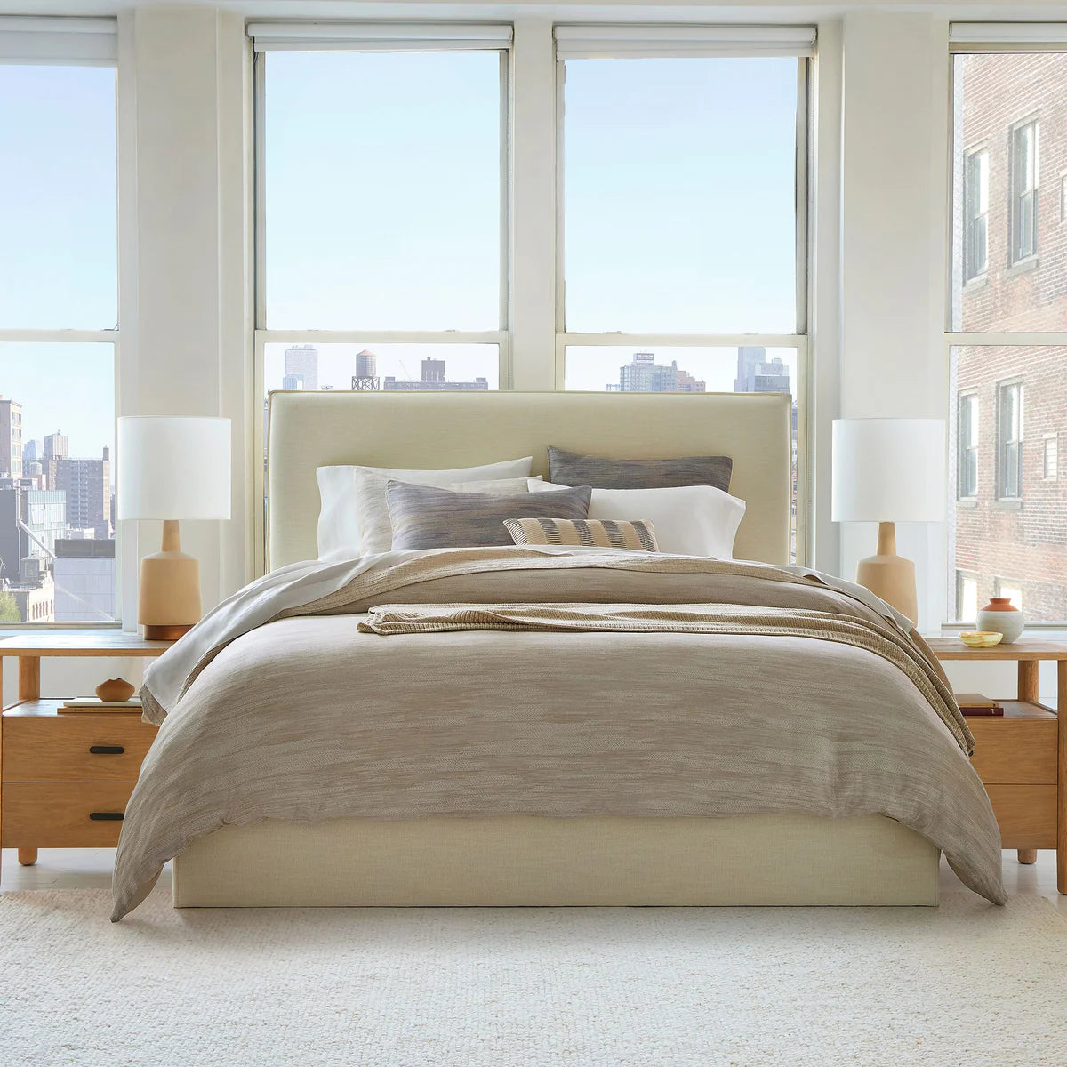 Neatly made bed with beige bedding in a bright bedroom with large windows.
