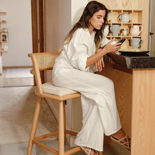 Woman in a white outfit sitting on a wooden chair in a kitchen, using a phone.